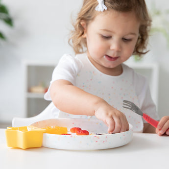 A toddler in a white shirt enjoys food from a Bumkins Silicone Grip Plate: Vanilla Sprinkle using a fork with a yellow cup nearby.