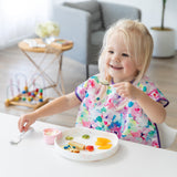 A smiling young girl enjoys dessert from a Bumkins Silicone Grip Plate: Vanilla Sprinkle with a blurred toy and chair in the background.