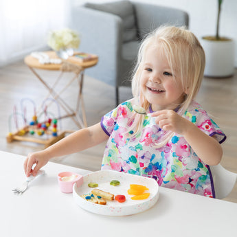 A smiling young girl enjoys dessert from a Bumkins Silicone Grip Plate: Vanilla Sprinkle with a blurred toy and chair in the background.