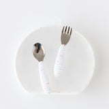 The Bumkins Grip Plate Essential 4-Piece Mealtime Set in Vanilla Sprinkle features a spoon, fork, and textured handles on a translucent plate.