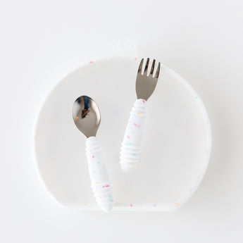 The Bumkins Grip Plate Essential 4-Piece Mealtime Set in Vanilla Sprinkle features a spoon, fork, and textured handles on a translucent plate.