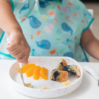 A child's hand uses a fork to eat food from a Bumkins Silicone Grip Plate: Vanilla Sprinkle, wearing a sea creature bib at a white table.