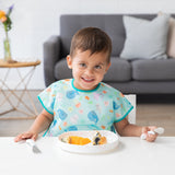 A smiling child sits at a white table wearing a colorful bib, ready to eat from a Bumkins Silicone Grip Plate: Vanilla Sprinkle.