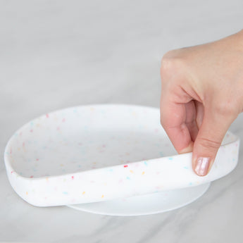 A hand holds a Bumkins Silicone Grip Plate: Vanilla Sprinkle, demonstrating the suction cup bottom on a marble surface.