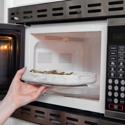 A hand placing a plate of food into a microwave oven. The microwave door is open, revealing a digital control panel on the side. The plate holds green beans. The hand has a ring on the ring finger.