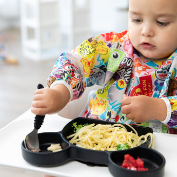 A toddler in a colorful bib enjoys spaghetti from Bumkins Disney Mickey Mouse feeding set at the table.
