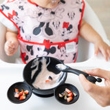 A baby in a Bumkins Growing with Mickey Mouse feeding set is guided by an adult hand as they enjoy yogurt and fruit.