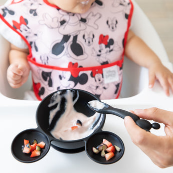 A baby in a Bumkins Growing with Mickey Mouse feeding set is guided by an adult hand as they enjoy yogurt and fruit.