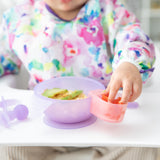 A child in a bib reaches for food from a Bumkins Silicone Little Dipper Combo 2 Pack: Magenta Jelly attached to a bowl on a high chair tray.
