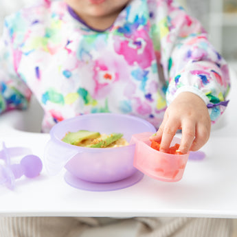 A child in a bib reaches for food from a Bumkins Silicone Little Dipper Combo 2 Pack: Magenta Jelly attached to a bowl on a high chair tray.