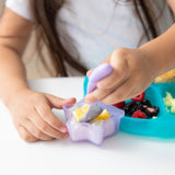 A child uses purple spoon for yogurt from a Bumkins Silicone Little Dipper Combo 2 Pack: Magenta Jelly next to a blue plate on a white table.
