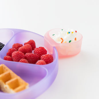 A Bumkins Silicone Little Dipper Combo 2 Pack: Magenta Jelly with whipped cream next to a plate of berries and waffles on a white background.