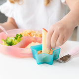 A child dips an apple slice into peanut butter from a Bumkins Silicone Little Dipper Combo 2 Pack: Aqua Jelly at a white table.