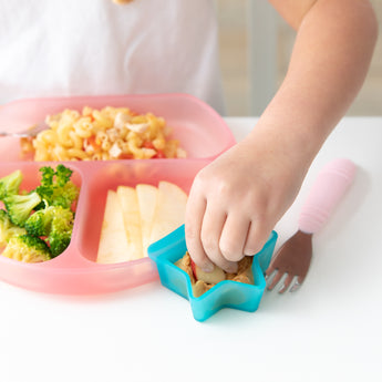 A child's hand reaches to dip an apple slice in peanut butter from a Bumkins Silicone Little Dipper Combo 2 Pack: Aqua Jelly at a white table.