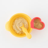 Top-down view of Bumkins Silicone First Feeding Set: Pineapple and a red heart-shaped cup filled with mashed food on a white background.