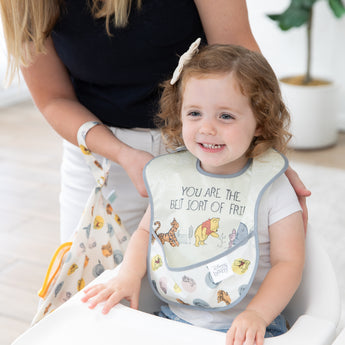 A woman holds Bumkins On-The-Go Mealtime 7-Piece Set: Pooh Bear + Friends as she adjusts a bib on a smiling young girl in a high chair.