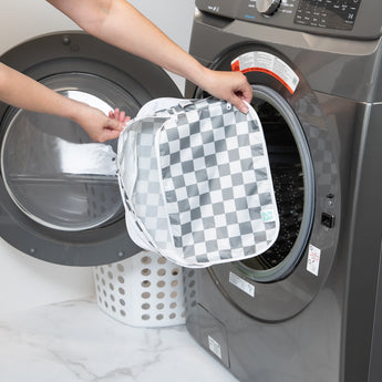 A person holds a bag from Bumkins The Outfit Organizer Essential 5-Piece Set: Charcoal Check in front of an open front-loading washing machine.