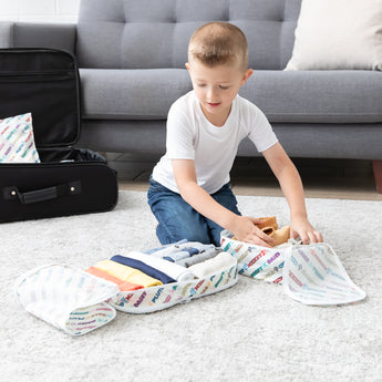 A young boy packs clothes into the Bumkins Packing Cube + Clear Travel Bag Set: Disney Mickey & Friends Varsity by his suitcase.