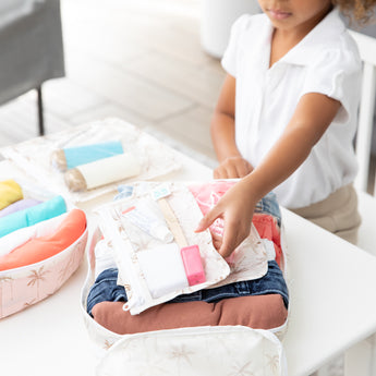 A child unpacks toiletries from a bag from Bumkins The Outfit Organizer Essential 5-Piece Set: Palm Daze on a white table.