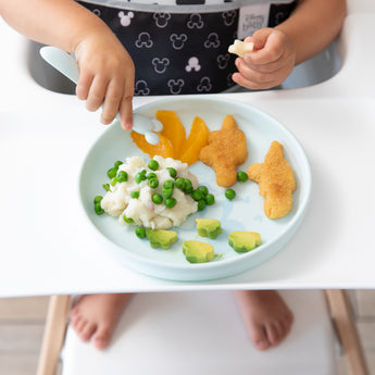 A child enjoys a meal of potatoes, peas, nuggets, peaches, and avocado from Bumkins Silicone Plate + Spoon Set: Disney Mickey Mouse.