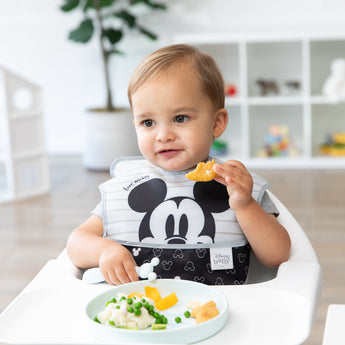 A toddler in a bib sits in a highchair, eating peas, mashed potatoes, and nuggets from Bumkins Silicone Plate + Spoon Set: Disney Mickey Mouse.