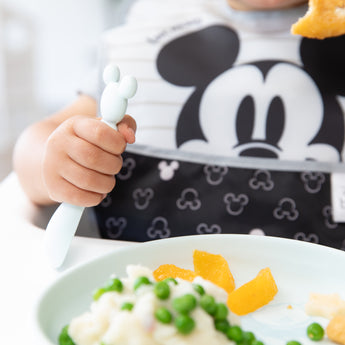 A child in a Mickey Mouse bib enjoying peas, oranges, and mashed potatoes from Bumkins Silicone Plate + Spoon Set: Disney Mickey Mouse.