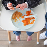A baby in a high chair wearing jeans and a bib delights in self-feeding with Bumkins Silicone Plate + Spoon Set: Disney Mickey Mouse.