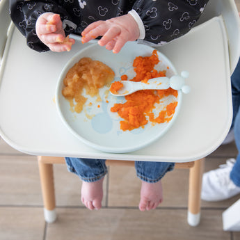 A baby in a high chair wearing jeans and a bib delights in self-feeding with Bumkins Silicone Plate + Spoon Set: Disney Mickey Mouse.