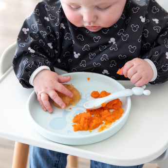 A baby in a high chair eats purees from Bumkins Silicone Plate + Spoon Set: Disney Mickey Mouse.