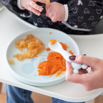 A baby in a high chair, wearing a bib, enjoys food from Bumkins Silicone Plate + Spoon Set: Disney Mickey Mouse with an adult's help.