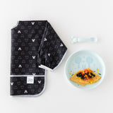 A black Mickey Mouse bib sits beside Bumkins Silicone Plate + Spoon Set: Disney Mickey Mouse filled with food on a white background.