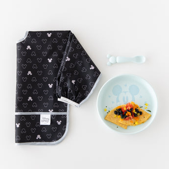 A black Mickey Mouse bib sits beside Bumkins Silicone Plate + Spoon Set: Disney Mickey Mouse filled with food on a white background.