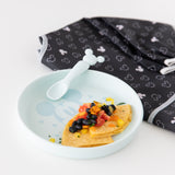 Bumkins Silicone Plate + Spoon Set: Disney Mickey Mouse filled with a tortilla, beans, and tomatoes sits next to a bib on a white background.