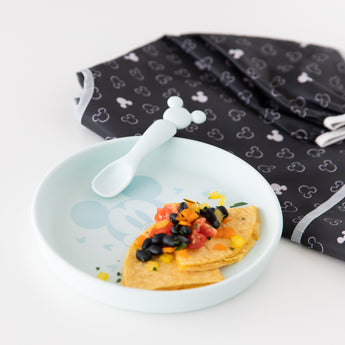 Bumkins Silicone Plate + Spoon Set: Disney Mickey Mouse filled with a tortilla, beans, and tomatoes sits next to a bib on a white background.