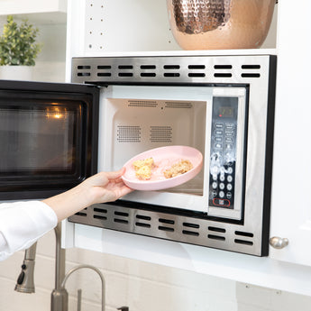 A person places a Bumkins Silicone Plate + Spoon Set: Disney Minnie Mouse filled with food into an open microwave.