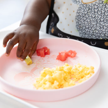 A toddlers hand reaches for food  from Bumkins Silicone Plate + Spoon Set: Disney Minnie Mouse with scrambled eggs, watermelon, and avocado.