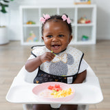 A smiling toddler with pink bows sits in a high chair using Bumkins Silicone Plate + Spoon Set: Disney Minnie Mouse to eat eggs and fruit.