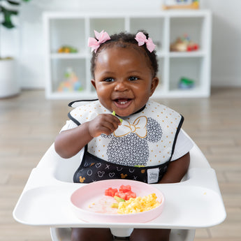 A smiling toddler with pink bows sits in a high chair using Bumkins Silicone Plate + Spoon Set: Disney Minnie Mouse to eat eggs and fruit.