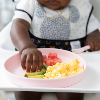 A baby in a high chair reaches for food from Bumkins Silicone Plate + Spoon Set: Disney Minnie Mouse with avocado, eggs, and watermelon.