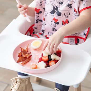 A child uses Bumkins Silicone Plate + Spoon Set: Disney Minnie Mouse to eat fruit, bacon, and an egg on a white high chair.