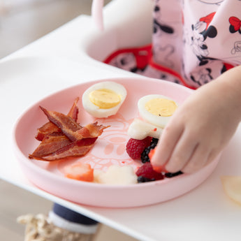 A child's hand reaches for a fruit from Bumkins Silicone Plate + Spoon Set: Disney Minnie Mouse filled with bacon, eggs, and berries.
