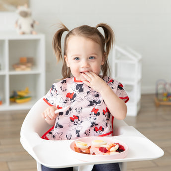 A toddler with pigtails in a high chair enjoys fruit from Bumkins Silicone Plate + Spoon Set: Disney Minnie Mouse with a playful expression.