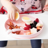 A child uses Bumkins Silicone Plate + Spoon Set: Disney Minnie Mouse to enjoy breakfast, wearing a Disney bib in a white high chair.