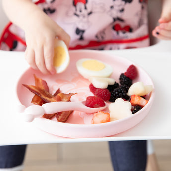 A child uses Bumkins Silicone Plate + Spoon Set: Disney Minnie Mouse to enjoy breakfast, wearing a Disney bib in a white high chair.
