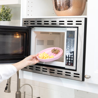 A hand places Bumkins Silicone Plate + Spoon Set: Disney Princess with eggs and sausage into an open microwave.