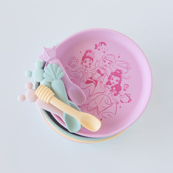 Top down view of Bumkins Silicone Plate + Spoon Set: Disney Princess, stacked on top of other similar plates on a white background.