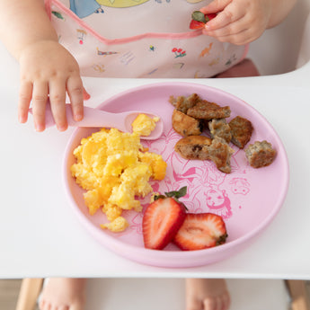 A child in a high chair enjoys breakfast from Bumkins Silicone Plate + Spoon Set: Disney Princess while wearing a pink bib.
