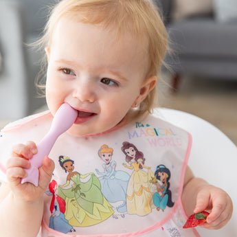 A toddler in a high chair holds a spoon from Bumkins Silicone Plate + Spoon Set: Disney Princess in their mouth while holding a strawberry.