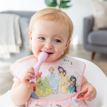 A smiling blonde baby in a bright room holds a spoon from Bumkins Silicone Plate + Spoon Set: Disney Princess, wearing a princess bib.