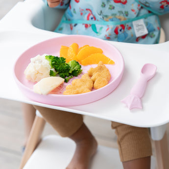 A child in a high chair with Bumkins Silicone Plate + Spoon Set: Disney Princess holding broccoli, potatoes, orange slices, and chicken nuggets.